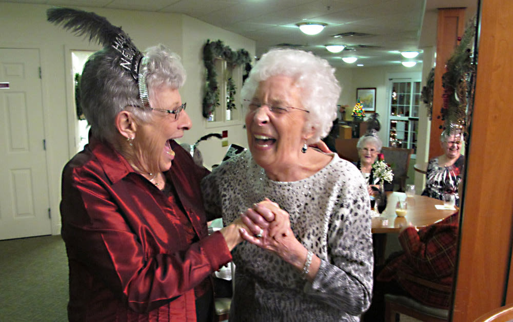Two elderly women laughing and holding hands indoors